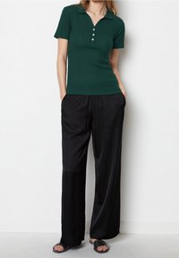 Kobieta w ciemnozielonej, krótkiej koszulce polo z ribby, czarnych spodniach o szerokich nogawkach, z rękami w kieszeniach i czarnych klapkach z odkrytymi palcami.