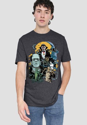Young man wearing dark gray shirt with colorful retro horror movie characters including Frankenstein, Dracula, and a mummy on front.