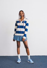 Jeune femme avec de longues tresses portant une chemise à rayures bleues et blanches à manches longues, une jupe en denim, des chaussettes blanches et des baskets bleues, se tenant sur un tapis bleu.
