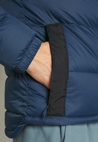 Blue puffer jacket with a textured surface, featuring a black zippered pocket and elastic cuffs. Smooth fabric with a subtle grid pattern.