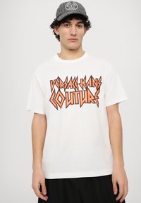 Tricou alb din bumbac, decorat cu un grafic îndrăzneț și unghiular în nuanțe de portocaliu și negru cu textul "VERSACE JEANS COUTURE". Modelul poartă o șapcă gri cu logo.