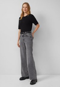 Frau in einem schwarzen kurzärmeligen Oberteil, hoch taillierten grauen weiten Jeans mit einem schwarzen Gürtel und schwarzen klobigen Stiefeln, steht vor einer schlichten Wand.