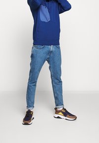 Felpa navy con tasca a marsupio, jeans azzurri chiari e sneakers marroni e navy con accenti gialli. Le sneakers hanno un design chunky e suole testurizzate.