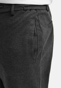 Close-up van donkergrijze, gestructureerde broek met zijzak en tailleband, gedragen met een wit topje dat zichtbaar is bij de taille.