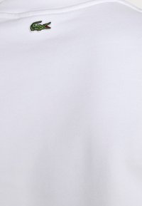 Polo blanc en coton avec un petit logo crocodile Lacoste vert brodé sur le côté gauche du col. Texture lisse.