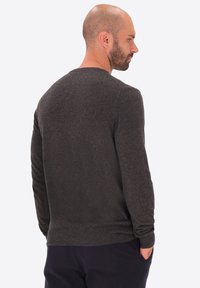 Uomo con barba corta e testa calva sta in posizione di profilo, indossando un maglione a maniche lunghe grigio scuro e pantaloni neri, con le mani nelle tasche.