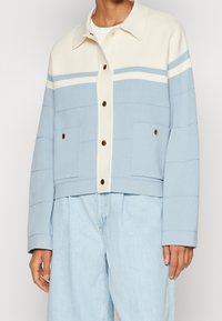 Jasnoniebieska krótka kurtka z kremowymi górnymi częściami, poziomymi paskami, złotymi guzikami i dwoma przednimi kieszeniami. Miękka tekstura tkaniny.