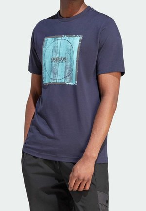Homme portant un t-shirt Adidas bleu marine avec un carré abstrait bleu clair et un texte noir, associé à un pantalon noir sur un fond uni.