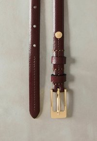 Bruine leren riem met een gladde textuur, voorzien van een gouden rechthoekige gesp en een decoratief hardwareaccent. Meerdere gaatjes voor verstelbaarheid.