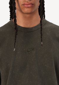 Tricou din bumbac de culoare verde măslin, având decolteu rotund și o textură ușor uzată. Logo-ul Nike, brodat, este vizibil și de aceeași culoare.
