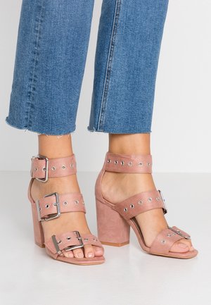 Różowe zamszowe sandały na wysokim obcasie z szerokimi paskami, klamrami, metalowymi akcentami i otwartymi palcami. Obcas jest masywny i fakturowany.