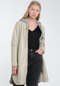 Beige nahkatakki harmaalla hupulla, jossa on vetoketju ja sivutaskut, päällä musta V-päähine ja mustat farkut.
