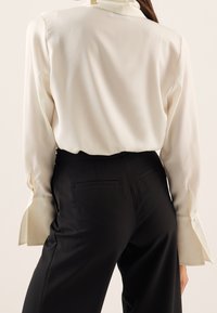 Weiße Seidenbluse mit langen Ärmeln und geknöpften Manschetten, kombiniert mit schwarzen, maßgeschneiderten Hosen mit glattem Finish und Gesäßtaschen.