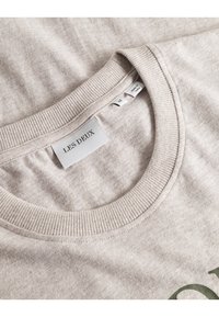 Lett taupe crewneck t-skjorte med ribbestrikket krage. Har "LES DEUX" merke og grønn trykt tekst. Bomullstoff med myk tekstur.