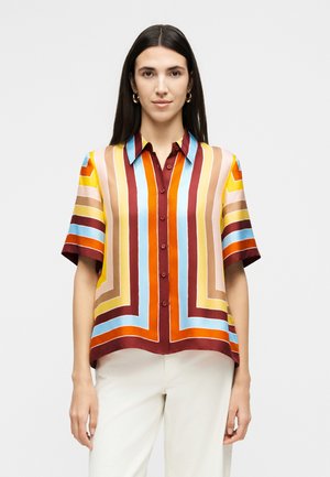 Kobieta o długich ciemnych włosach, ubrana w krótkim rękawem, zapinaną na guziki koszulę z wielokolorowymi pionowymi i poziomymi paskami oraz białe spodnie.