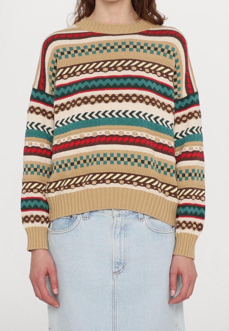 Femme portant un pull beige avec des motifs horizontaux multicolores et une jupe en jean bleu clair, debout sur un fond blanc.