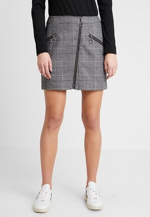 Kvinde iført en grå ternet mininederdel med lynlåsdetalje foran og sorte lynlåslommer, kombineret med hvide sneakers og en sort ribbet top.