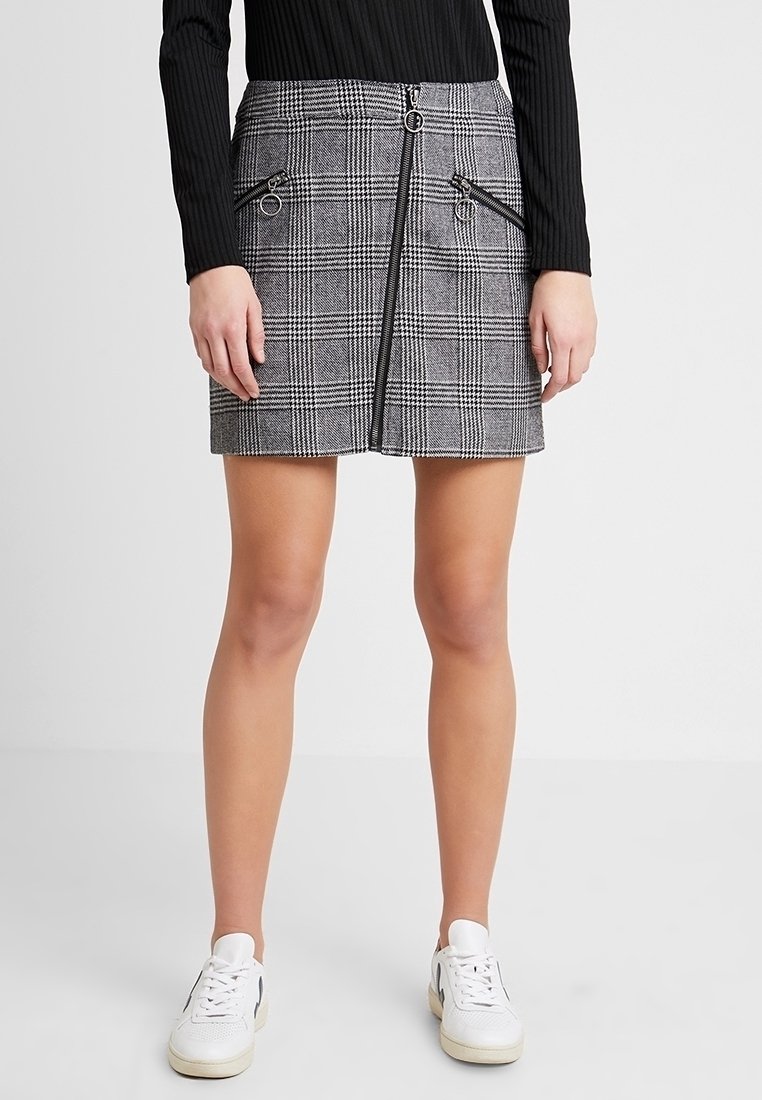 Femme portant une mini-jupe grise à carreaux avec un détail de fermeture éclair à l'avant et des poches zippées noires, associée à des baskets blanches et un haut côtelé noir.