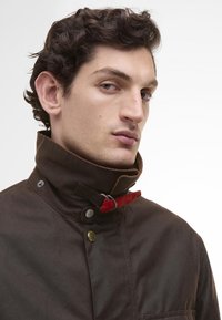 Barna viaszolt szövetdzseki magas gallérral, piros díszcsíkkal és patentos gombokkal a nyaknál. Simaszövet, minimális részletezéssel.