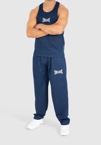 Marineblauwe sporttanktop en bijpassende joggers met ribbelstructuur en witte logo-accenten. Model draagt witte sportschoenen.