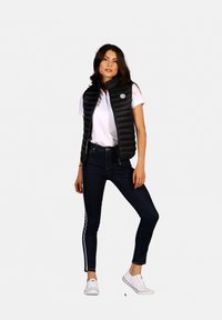 Gilet noir rembourré avec un col montant, associé à un polo blanc et un jean foncé avec des rayures latérales, complété par des baskets blanches.