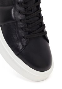 Zwarte leren sneaker met zwarte veters en dikke witte rubberen zool, met gedetailleerde textuur en stiksels op het bovenste voorste gedeelte.