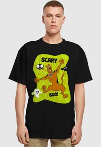 Melnā T-krekls ar krāsainu grafiku, kurā attēlots karikatūru suns "Scooby-Doo", apkārt kuram ir zirneklis, sikspārņi un spoks, uz zaļas tīkla fona.