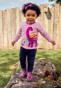 Gestreepte lange mouwen shirt met een paarse dinosaurus en bloemmotieven. G gedragen met een donkere spijkerbroek en paarse rubberen laarzen, staand op hout.