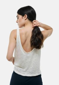 Haut blanc sans manches avec un dos décolleté, fabriqué dans un tissu léger et texturé. L'ourlet est légèrement courbé et il a une coupe décontractée.