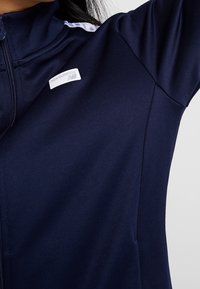 Námořnická bunda s plným zipem a vysokým límcem, s bílými vzorovanými detaily na ramenou a malým látkovým logem na hrudi.