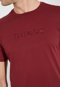 Hombre vistiendo una camiseta básica de cuello redondo color burdeos con el logotipo en relieve "GUESS" en el pecho, cortada por debajo de la nariz.