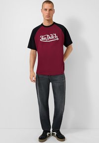 Bordeaux ir juodas raglan stiliaus marškinėliai su "Von Dutch" logotipu, derinami su tamsiomis džinsais ir juodais sportbačiais, stovintis prieš neutralų foną.