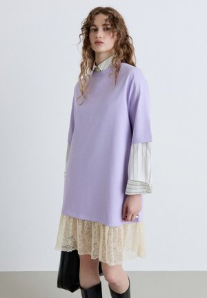 Giovane donna con capelli ricci indossa un vestito maglione lavanda sopra una camicia a righe e una gonna di pizzo, tiene una borsa nera, in piedi davanti a uno sfondo semplice.
