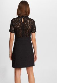 Zwarte jurk met een kant bodice, korte mouwen, een keyhole rugdetail en een getailleerde zwarte rok. De stof heeft een gestructureerd ontwerp.