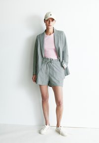 Hellgraue karierten Sakkos über einem rosa gerippten Top, kombiniert mit passenden grauen Shorts. Weiße Sneaker und eine beigen Cap runden das Outfit ab.