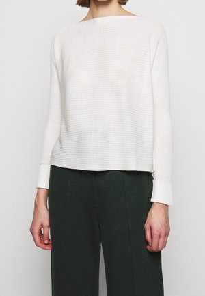 Personne portant un haut blanc côtelé à manches longues et un pantalon taille haute vert foncé, debout devant un fond uni.