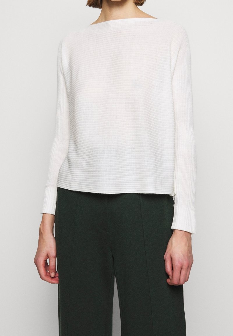 Personne portant un haut blanc côtelé à manches longues et un pantalon taille haute vert foncé, debout devant un fond uni.