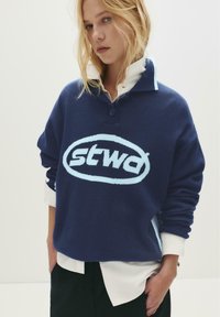 Blonde Frau, die einen marineblauen Pullover mit einem großen hellblauen "stwd"-Logo über einem weißen Hemd trägt, steht mit den Händen in den Taschen.