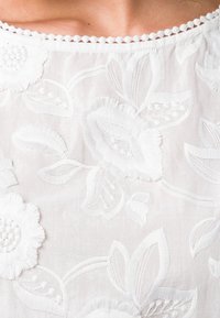 Tela blanca bordada con diseños florales que presentan pétalos y hojas texturizadas. El escote tiene un borde en forma de ondas.