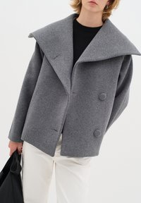 Grahamisch melierter Wollmischmantel mit großem Kragen und doppelreihiger Knopfverschluss, charakterisiert durch eine kastige Form und einen entspannten Schnitt. Begleitet von einer schwarzen Tasche.