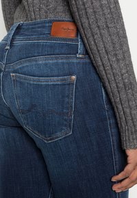 Blå denimjeans med en texturerad yta, som har en brun läderlogotyp, samt två bakfickor med sydda detaljer.