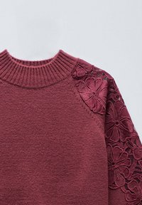 Jersey burdeos con cuello redondo acanalado y bordado de encaje floral en el hombro derecho y la manga, sobre un fondo liso.