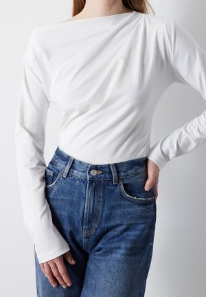 Osoba ubrana w dopasowaną białą bluzkę z długim rękawem wpuszczoną w niebieskie jeansy z wysokim stanem, z jedną ręką na biodrze, a drugą swobodnie luźną.