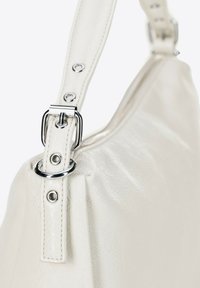 Kremowa skórzana torebka o teksturowanej powierzchni, z metalowym zapięciem i okrągłymi akcentami na regulowanym pasku.