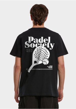 Person som har på seg en svart T-skjorte med stor hvit tekst "Padel Society" og grafikk av en padelracket og ball på baksiden.