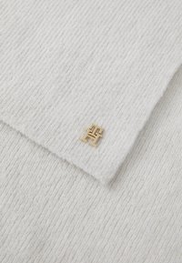 White ribbed sweater fabric featuring a small gold logo accent in the corner, highlighting a soft texture and clean design.