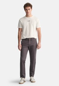 Witte katoenen T-shirt met zwarte tekst, gecombineerd met donkergrijze katoenen broek en witte sneakers met zwarte accenten. Model staat tegen een lichte achtergrond.