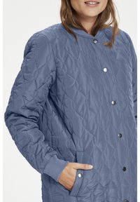 Gesteppte blaue Jacke mit Stehkragen, Druckknöpfen und seitlichen Taschen. Mittels einer strukturierten Rautenoptik und gerippten Bündchen versehen.