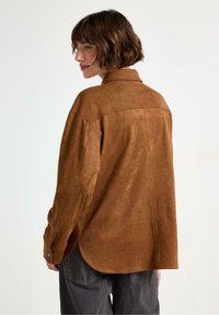 Camicia in suede marrone con colletto, maniche lunghe e orlo curvo. Presenta polsini con bottoni e una texture liscia e morbida.