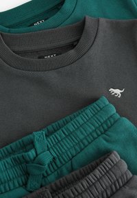 Twee sweatshirts in donkergrijs en teal, met ribgebreide halslijnen en een klein geborduurd ontwerp. Daaronder, drie paren tapered sweatpants in teal en grijs.